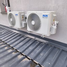 DG ALLINSTAL Dominik Goleń - Dwa białe klimatyzatory AUX DC Inverter zamontowane na szarej ścianie nad metalowym dachem. Widok z dołu, lekko z prawej strony. Na parapecie wiadro i narzędzia.