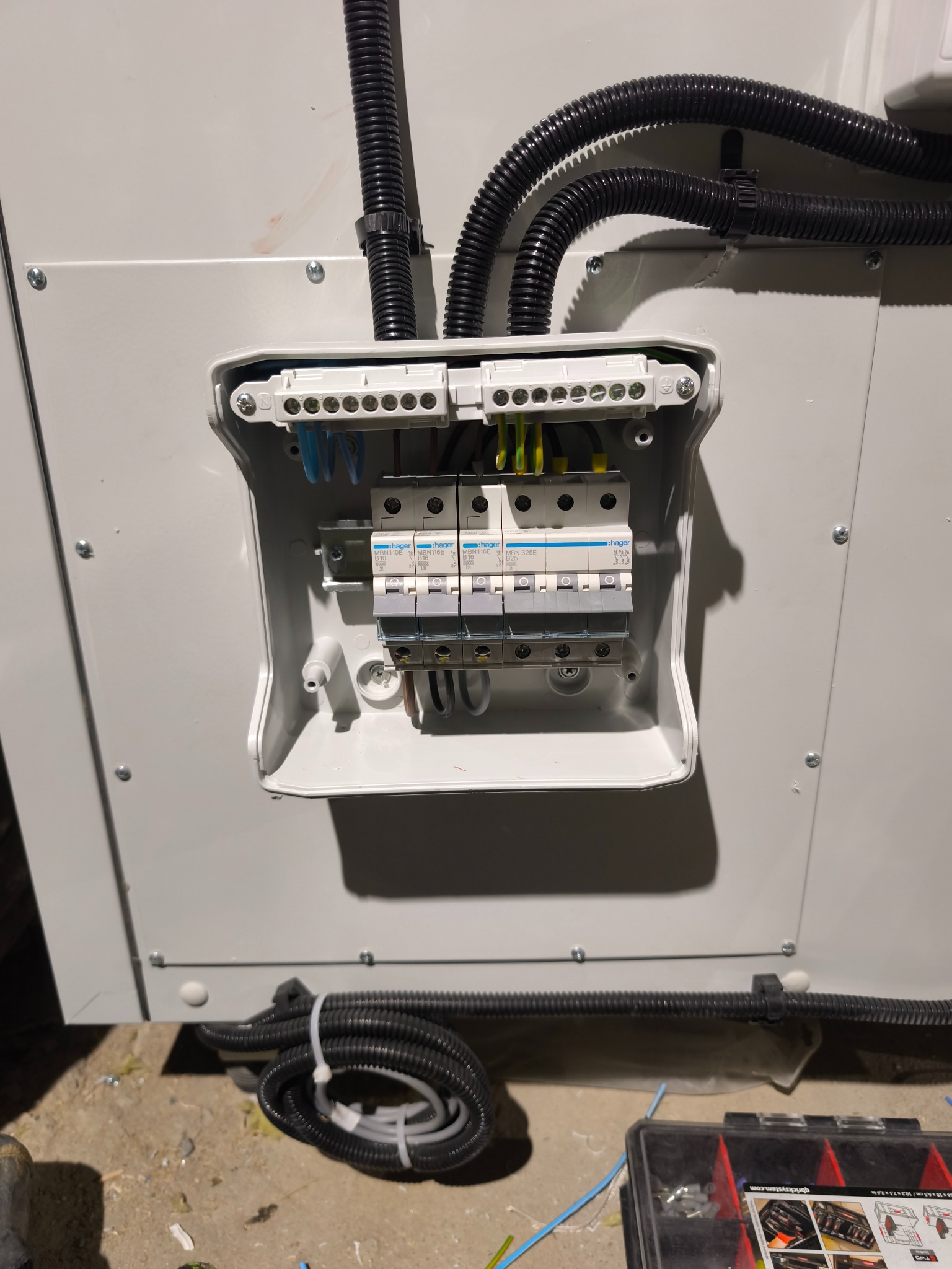 Rozdzielnica elektryczna z bezpiecznikami Hager w białej obudowie, widoczne kable w peszlach i narzędzia, instalacja w trakcie montażu.