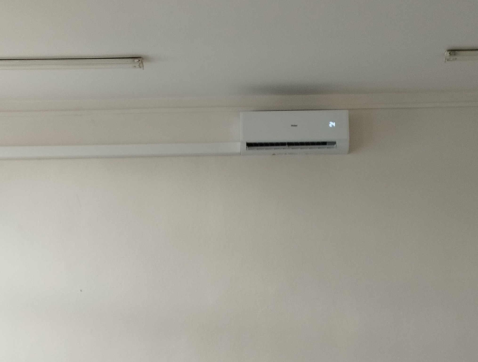 Biały klimatyzator Haier z wyświetlaczem temperatury 24°C zamontowany na jasnej ścianie, widoczne oświetlenie sufitowe. Minimalistyczny design.