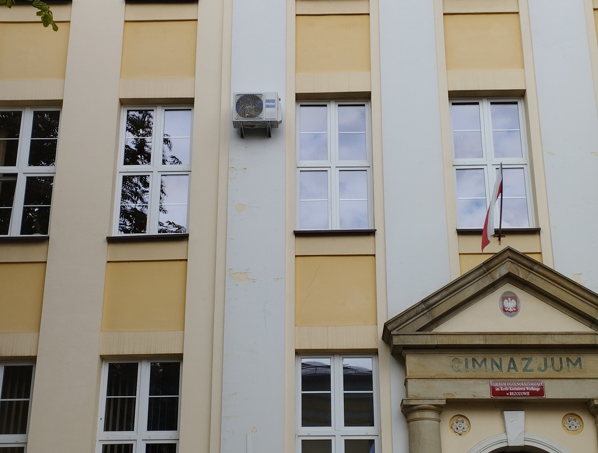 Elewacja budynku z oknami i zamontowanym klimatyzatorem Haier. Widoczne detale architektoniczne i flaga Polski. Kremowo-żółta kolorystyka.