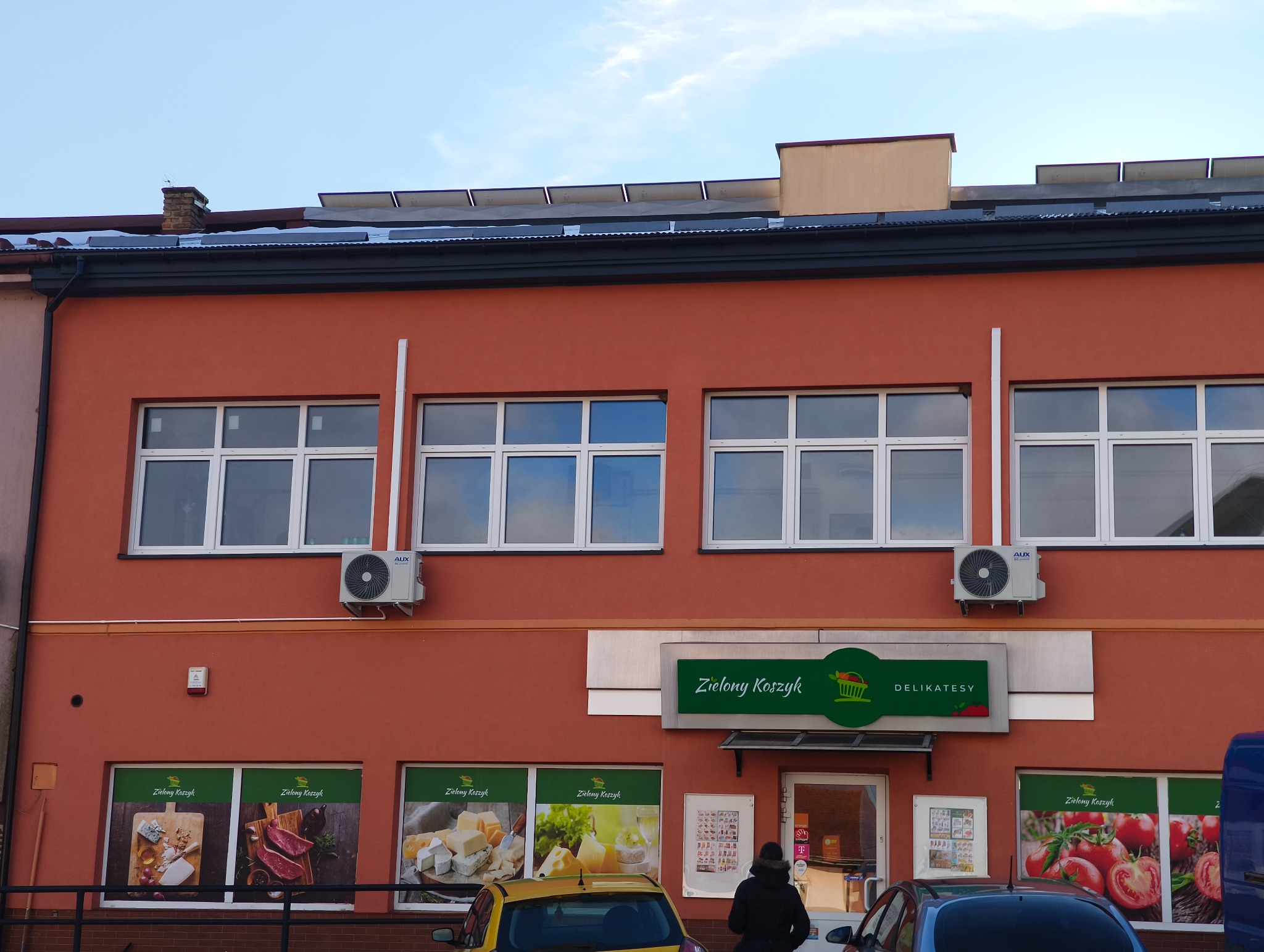 Czerwony budynek sklepu z klimatyzacją AUX pod oknami. Na dachu panele słoneczne. Widoczne okna z białymi ramami i szyld sklepu spożywczego.