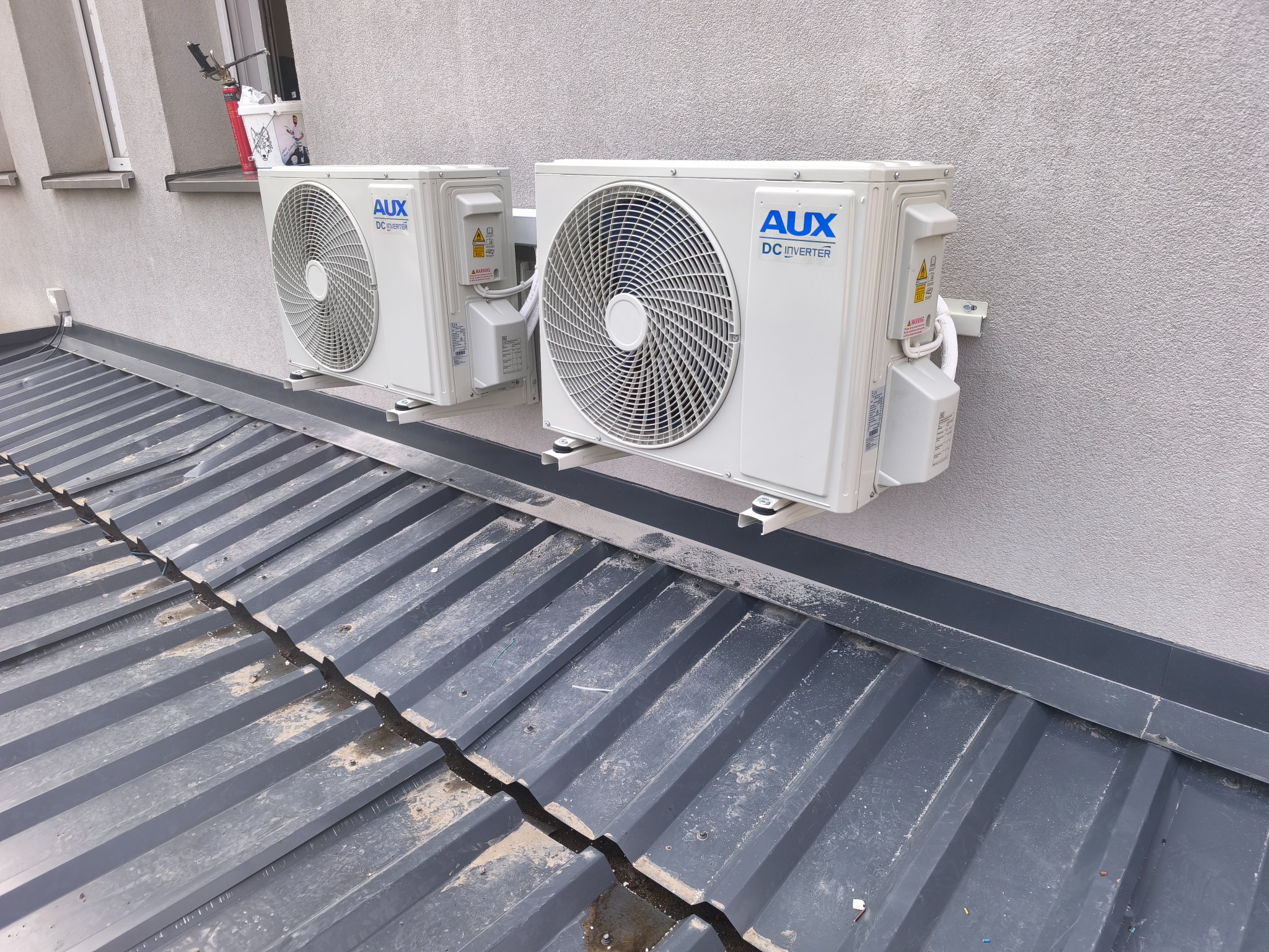 Dwa białe klimatyzatory AUX DC Inverter zamontowane na szarej ścianie nad metalowym dachem. Widok z dołu, lekko z prawej strony. Na parapecie wiadro i narzędzia.