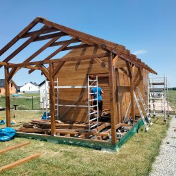 MAŁY-BUD USŁUGI REMONTOWO-BUDOWLANE PIOTR BYDŁOWSKI - Konstrukcja drewniana w trakcie budowy, widoczne rusztowania i narzędzia. Elementy domu szkieletowego na etapie montażu ścian i dachu, w tle zabudowa jednorodzinna.