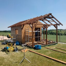 MAŁY-BUD USŁUGI REMONTOWO-BUDOWLANE PIOTR BYDŁOWSKI - Drewniany domek letniskowy w trakcie budowy, widoczne narzędzia i elementy konstrukcyjne na tle zielonego krajobrazu. Uwagę przykuwa precyzja wykonania szkieletu.