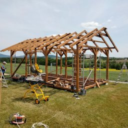 MAŁY-BUD USŁUGI REMONTOWO-BUDOWLANE PIOTR BYDŁOWSKI - Konstrukcja szkieletowa domu w trakcie budowy, widok z zewnątrz na drewniany szkielet z elementami dachu, narzędzia budowlane i drabiny na trawie, krajobraz w tle.