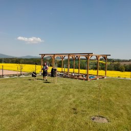 MAŁY-BUD USŁUGI REMONTOWO-BUDOWLANE PIOTR BYDŁOWSKI - Drewniana konstrukcja altany w trakcie budowy na zielonym trawniku, z widocznym pracownikiem i rozległym polem rzepaku w tle, w słoneczny dzień.