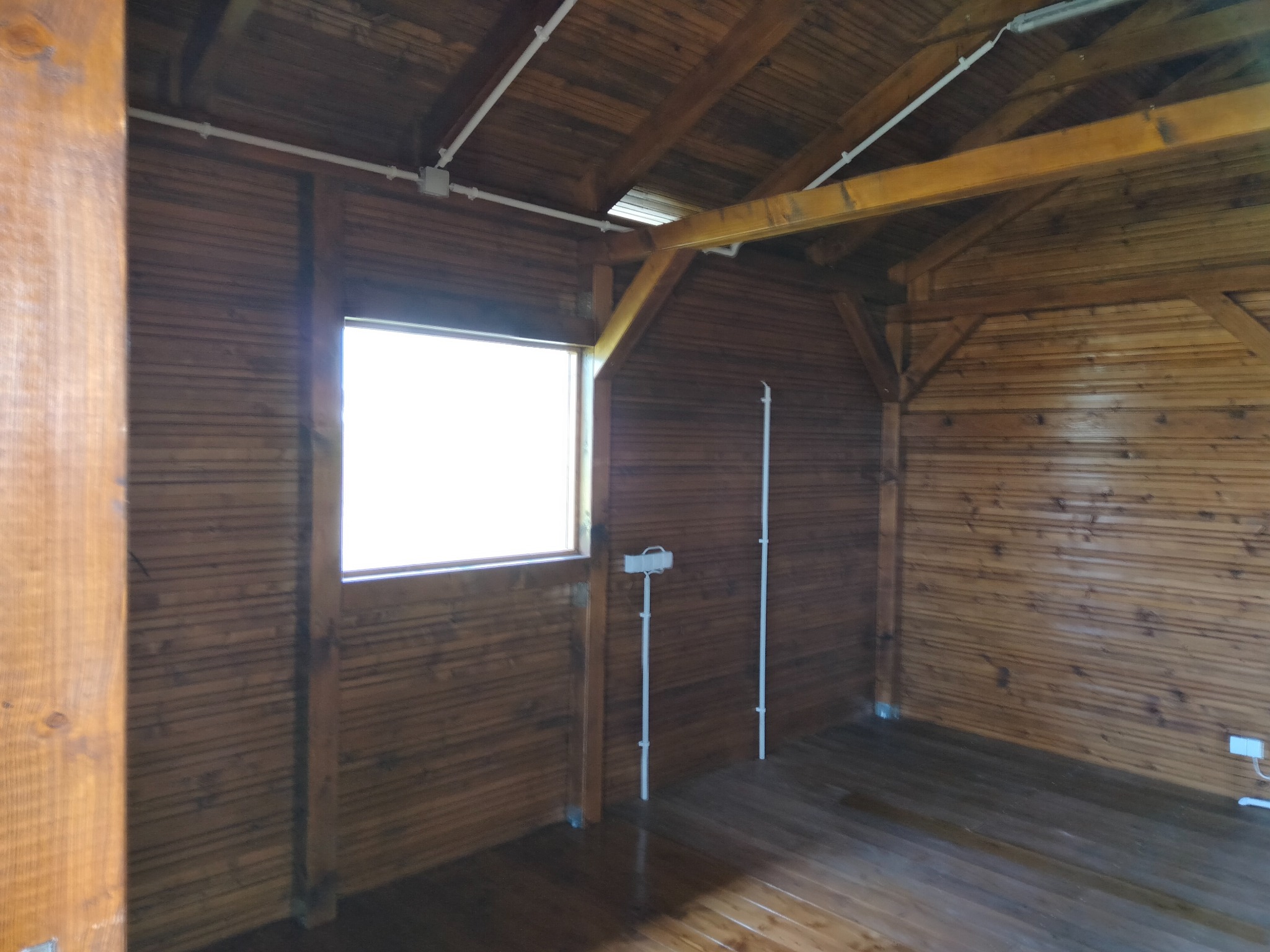 Wnętrze drewnianej konstrukcji w Świebodzicach z oknem i instalacją elektryczną. Widoczne deski na ścianach, podłodze i suficie. Ujęcie z perspektywy wnętrza.