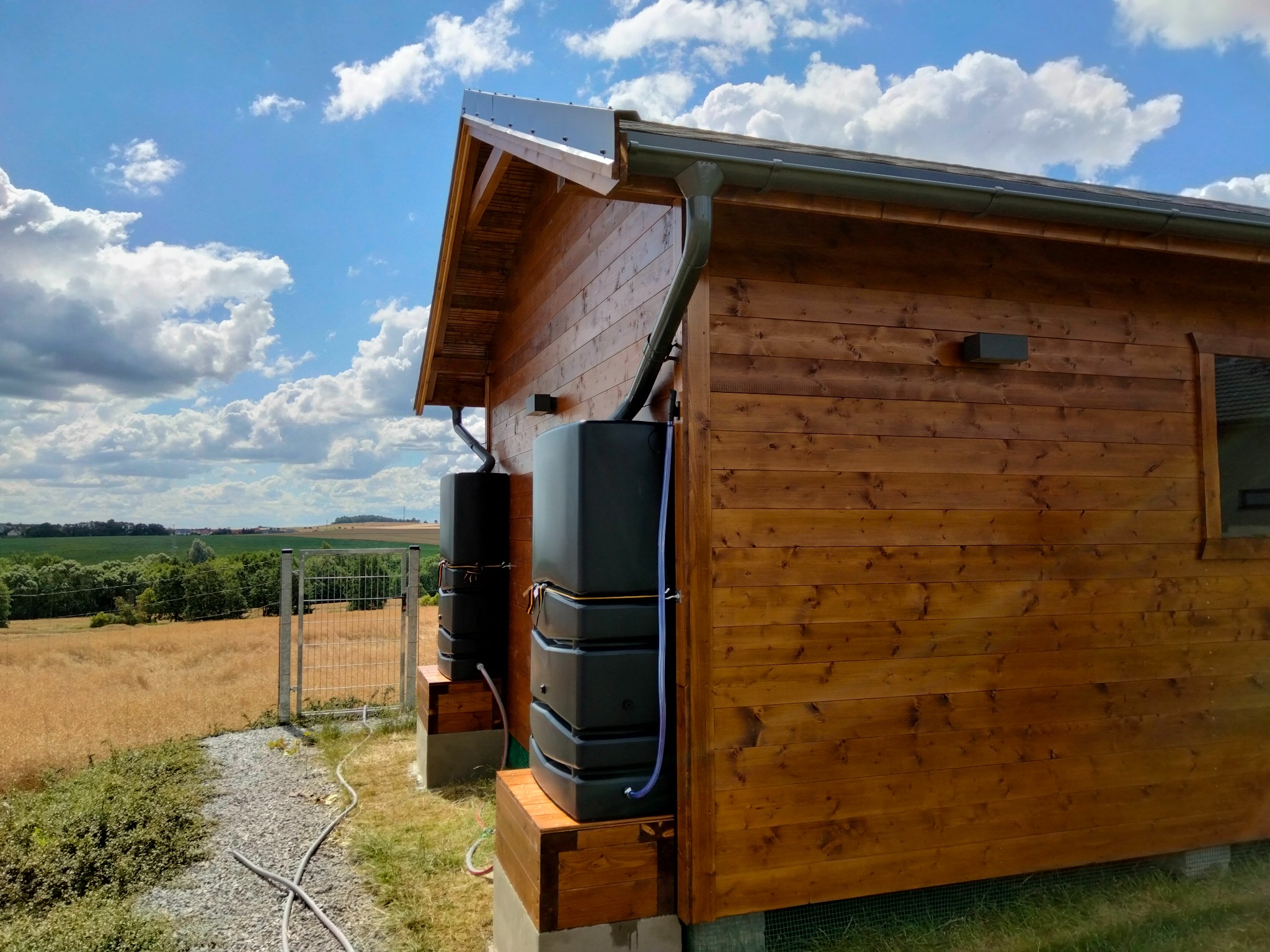 Drewniany dom szkieletowy z systemem zbierania deszczówki: zbiorniki na podwyższeniu, rynny i instalacja. W tle pola i błękitne niebo z chmurami.