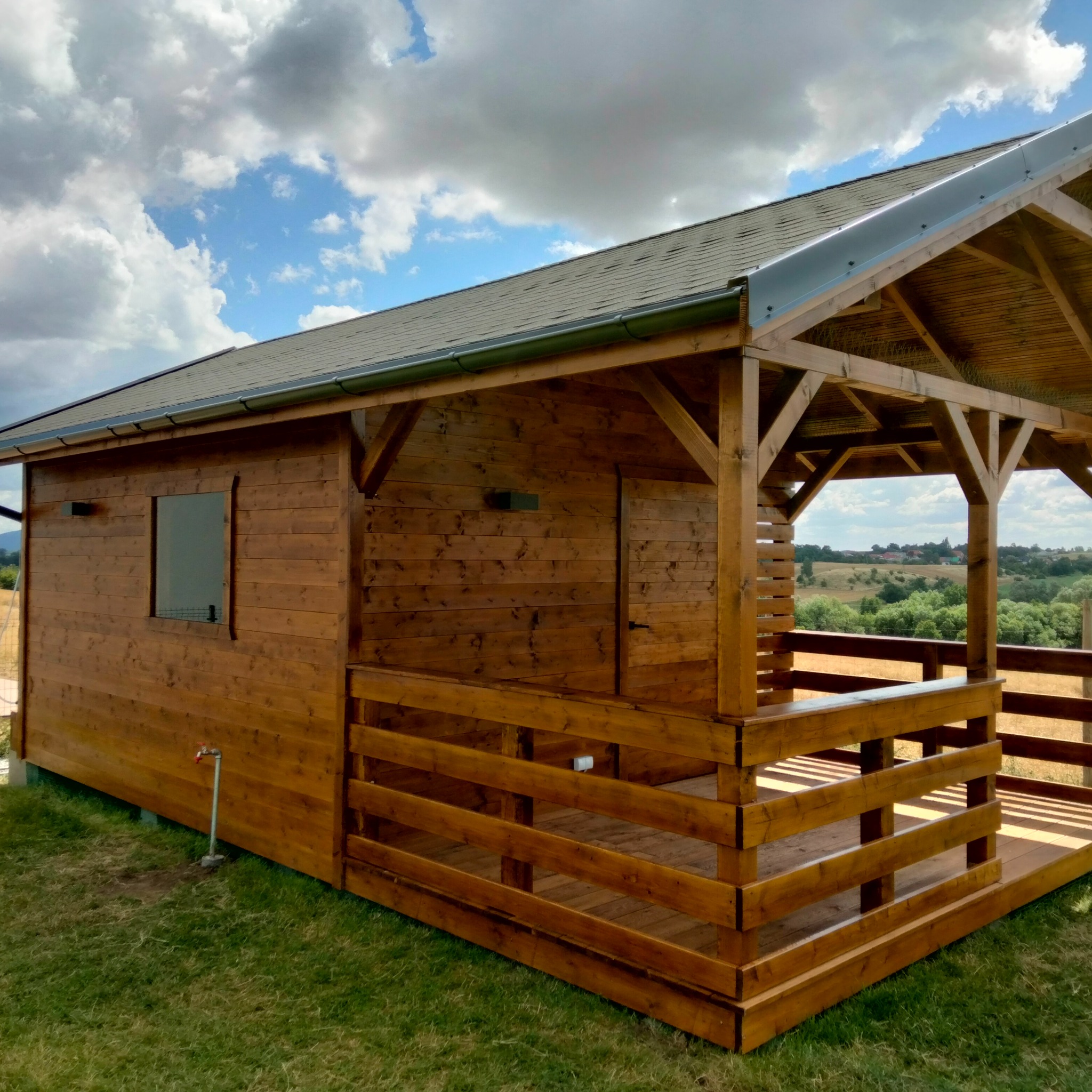 Drewniany domek letniskowy z zadaszonym tarasem, wykończony na brązowo, na tle zielonej trawy i błękitnego nieba z chmurami. Widoczny kran na zewnątrz.