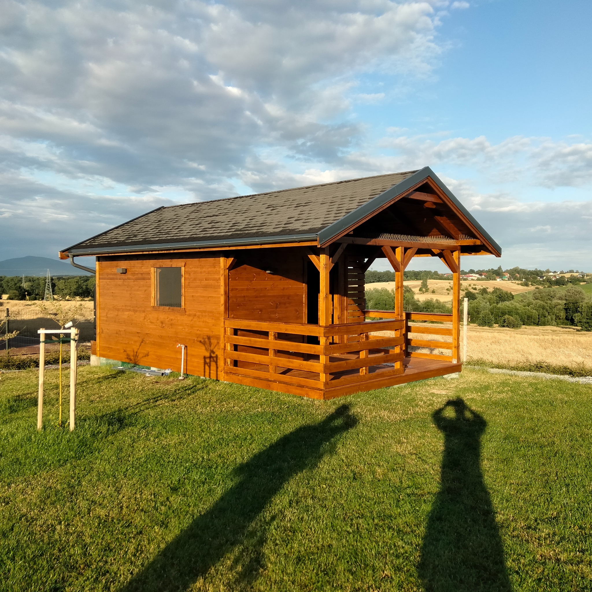 Drewniany domek letniskowy z zadaszonym tarasem, brązowe deski, ciemny dach, na zielonej trawie, w tle pola i wzgórza, cień osoby robiącej zdjęcie.
