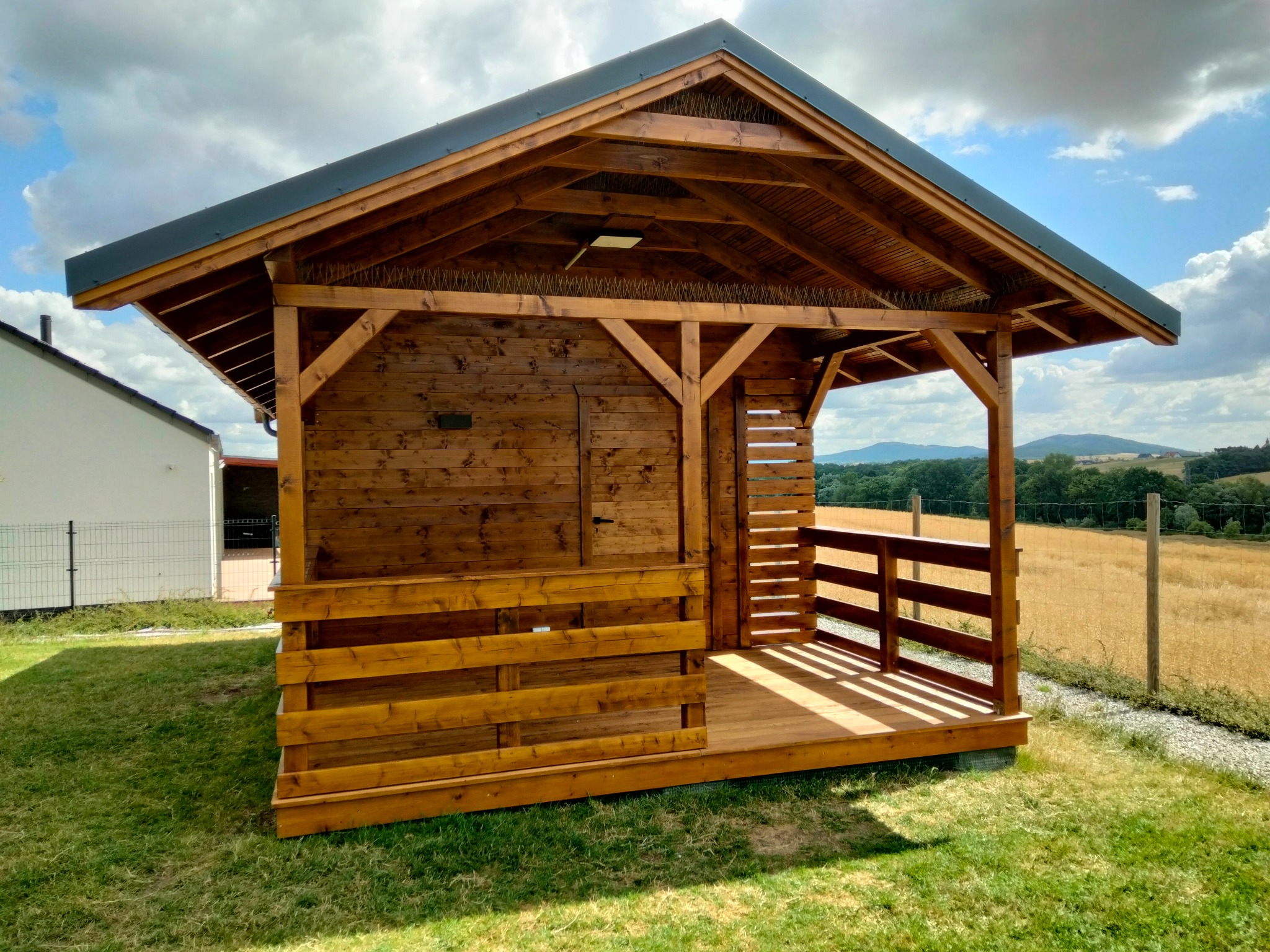 Drewniana altana z zamykaną częścią i otwartym tarasem, pomalowana na brązowo, na tle zielonego trawnika i krajobrazu z polami i wzgórzami.