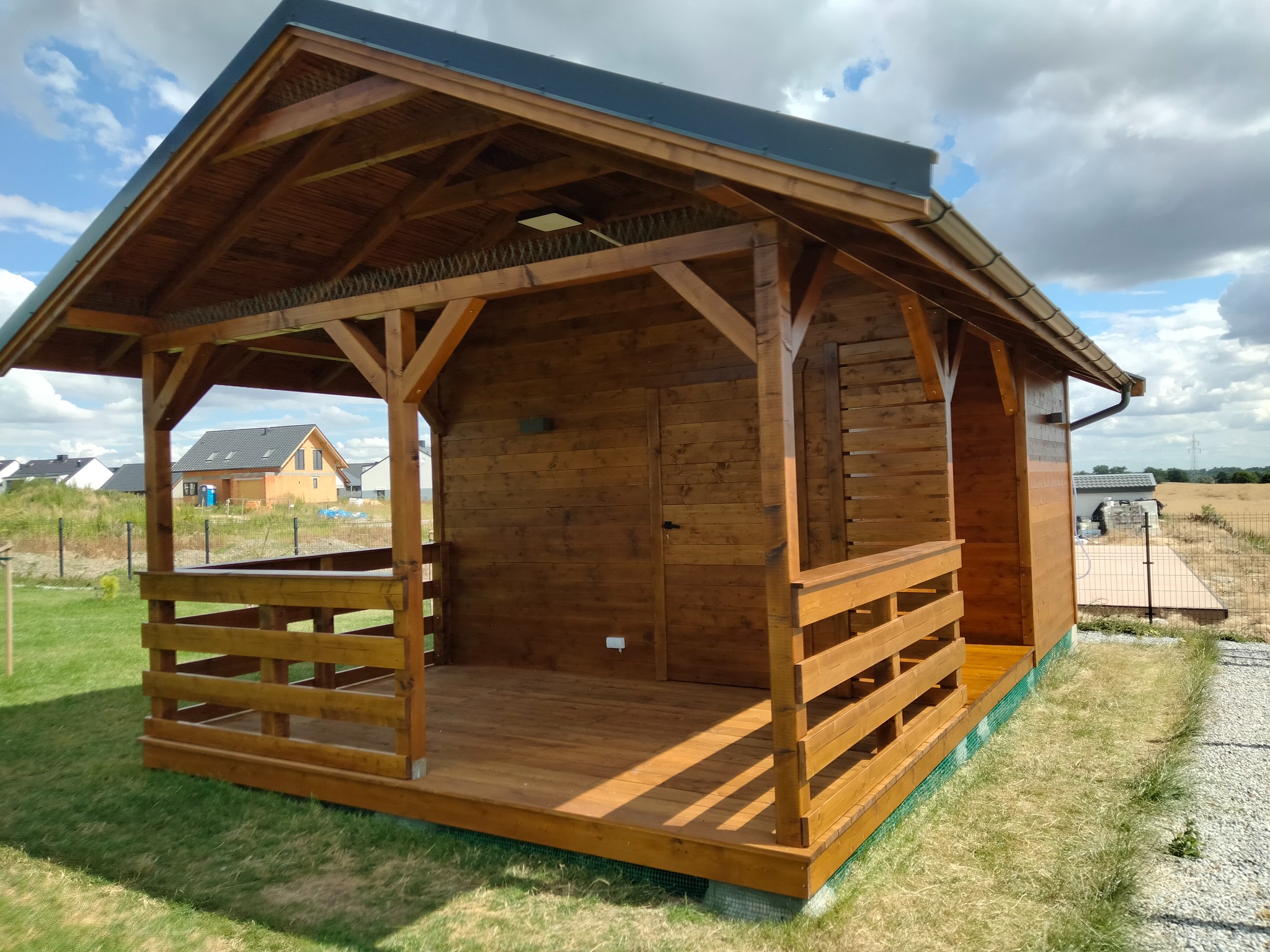 Drewniana altana z balustradą, wykończona na brązowo, na tle zielonej trawy i błękitnego nieba z chmurami. W tle widoczny dom jednorodzinny.