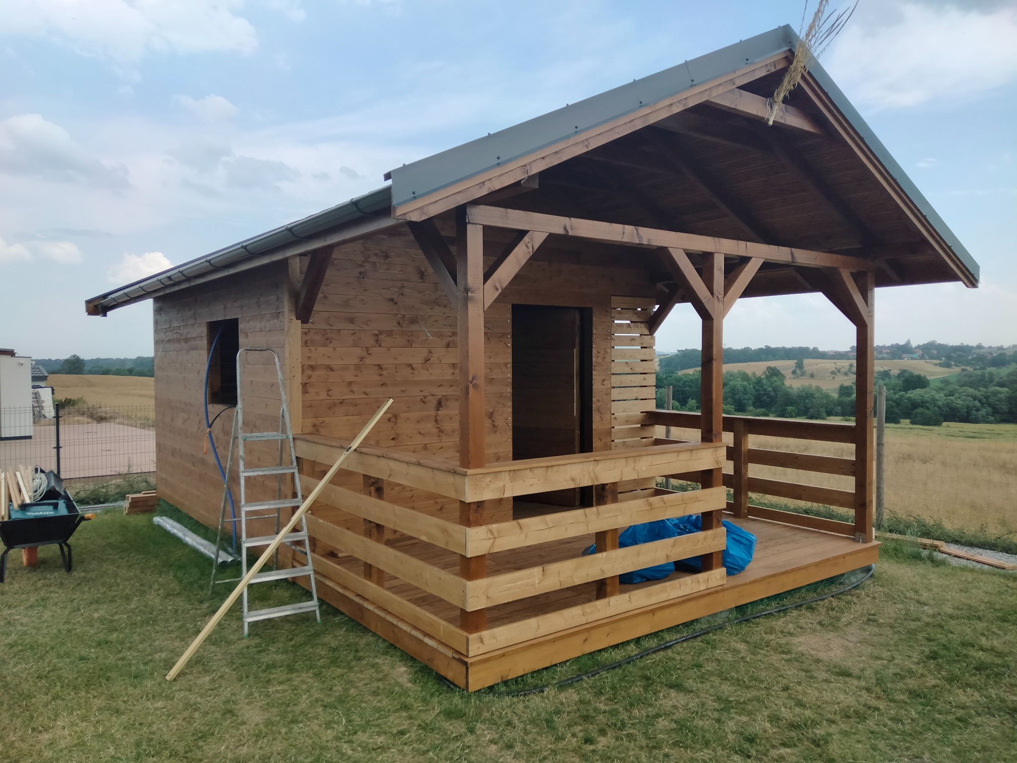 Drewniany domek letniskowy z zadaszonym tarasem w trakcie budowy, widoczna drabina i narzędzia, na tle zielonego krajobrazu.