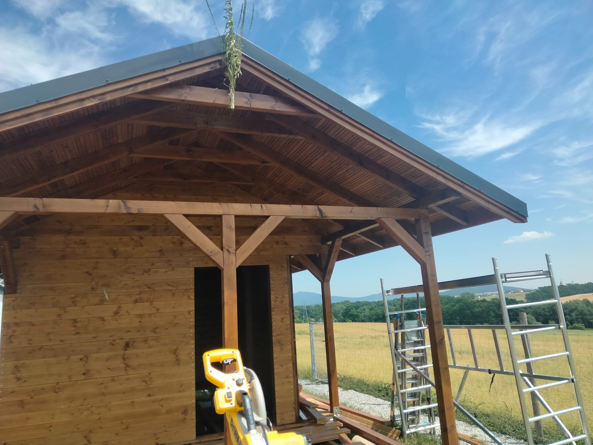 Drewniana konstrukcja altany w trakcie budowy, widoczne rusztowanie i narzędzie. W tle krajobraz polny pod błękitnym niebem.