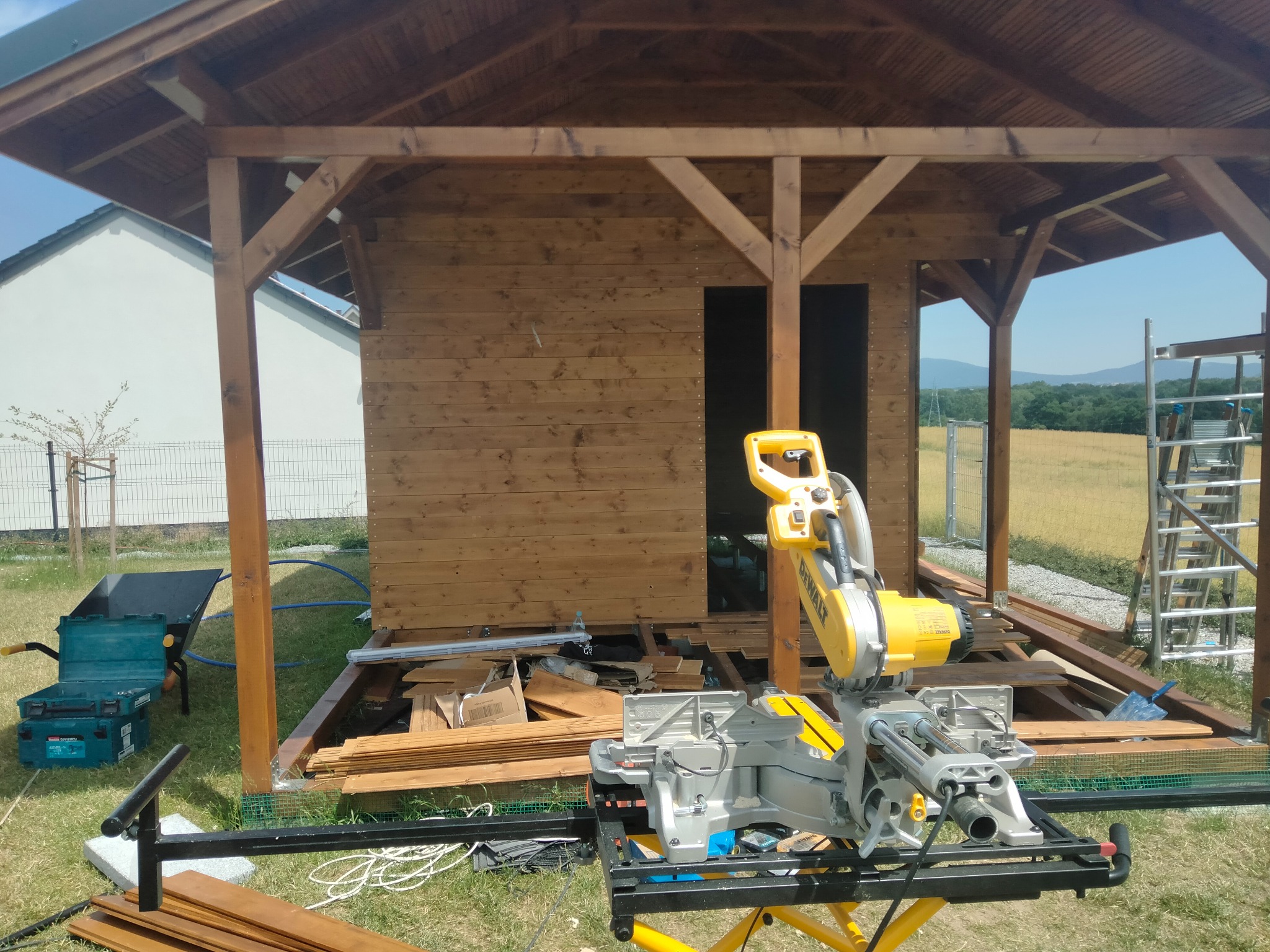 Budowa drewnianej altany: widoczna piła ukośnica DeWalt, elementy konstrukcyjne, narzędzia i częściowo ukończona podłoga. W tle krajobraz.