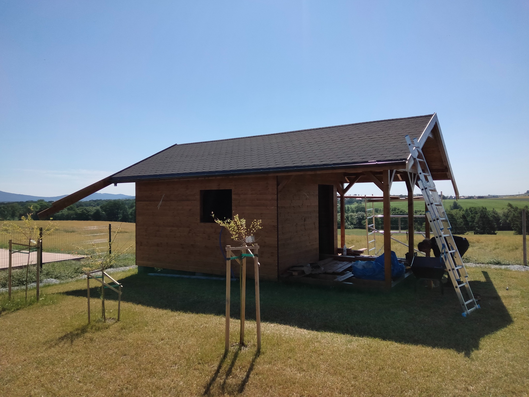 Drewniany domek z ciemnym dachem w trakcie budowy, z widoczną drabiną i elementami konstrukcyjnymi, na tle zielonego krajobrazu i błękitnego nieba.