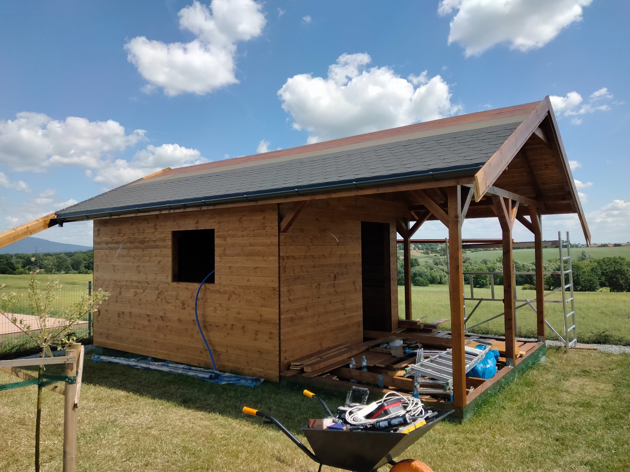Drewniany domek letniskowy w trakcie budowy, widok z zewnątrz, narzędzia w taczce, błękitne niebo z chmurami w tle. Widoczna konstrukcja dachu i ściany z desek.