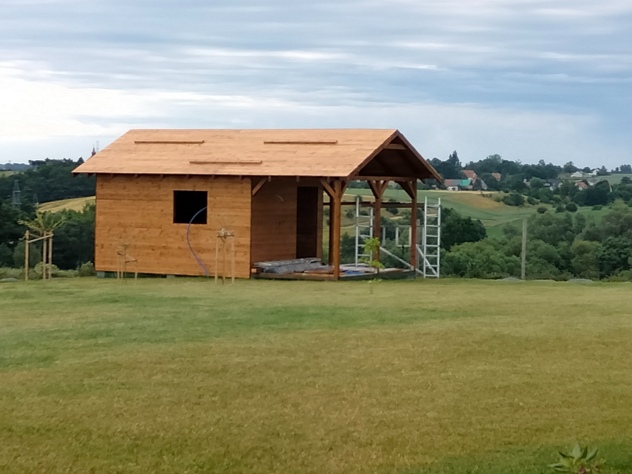 Drewniany domek letniskowy w trakcie budowy, z widoczną konstrukcją dachu i elementami szkieletowymi, na tle zielonego wzgórza i pochmurnego nieba.