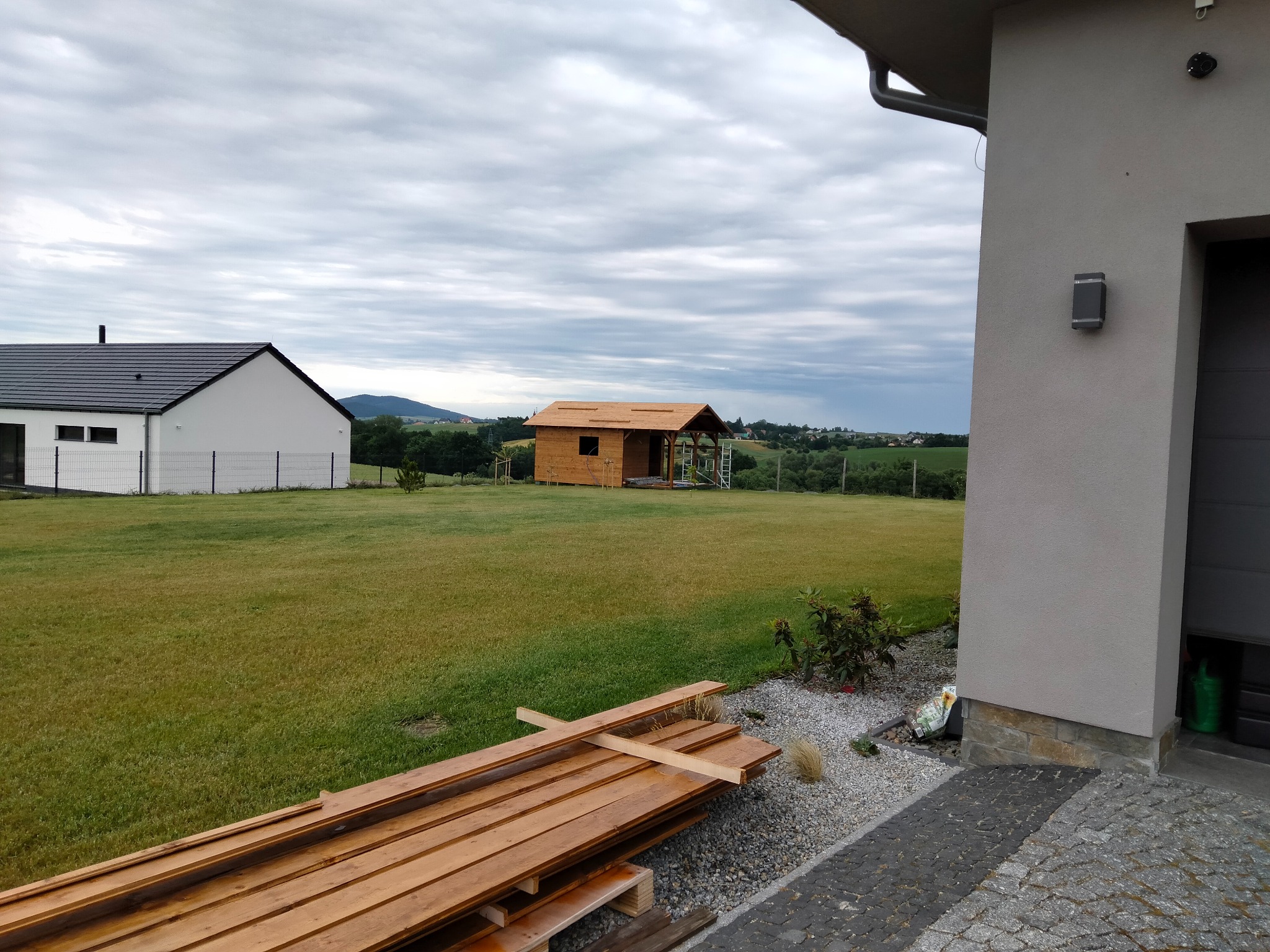 Budowa domku letniskowego na działce z widokiem. W tle dom jednorodzinny z białym ogrodzeniem. Na pierwszym planie deski i fragment elewacji domu.