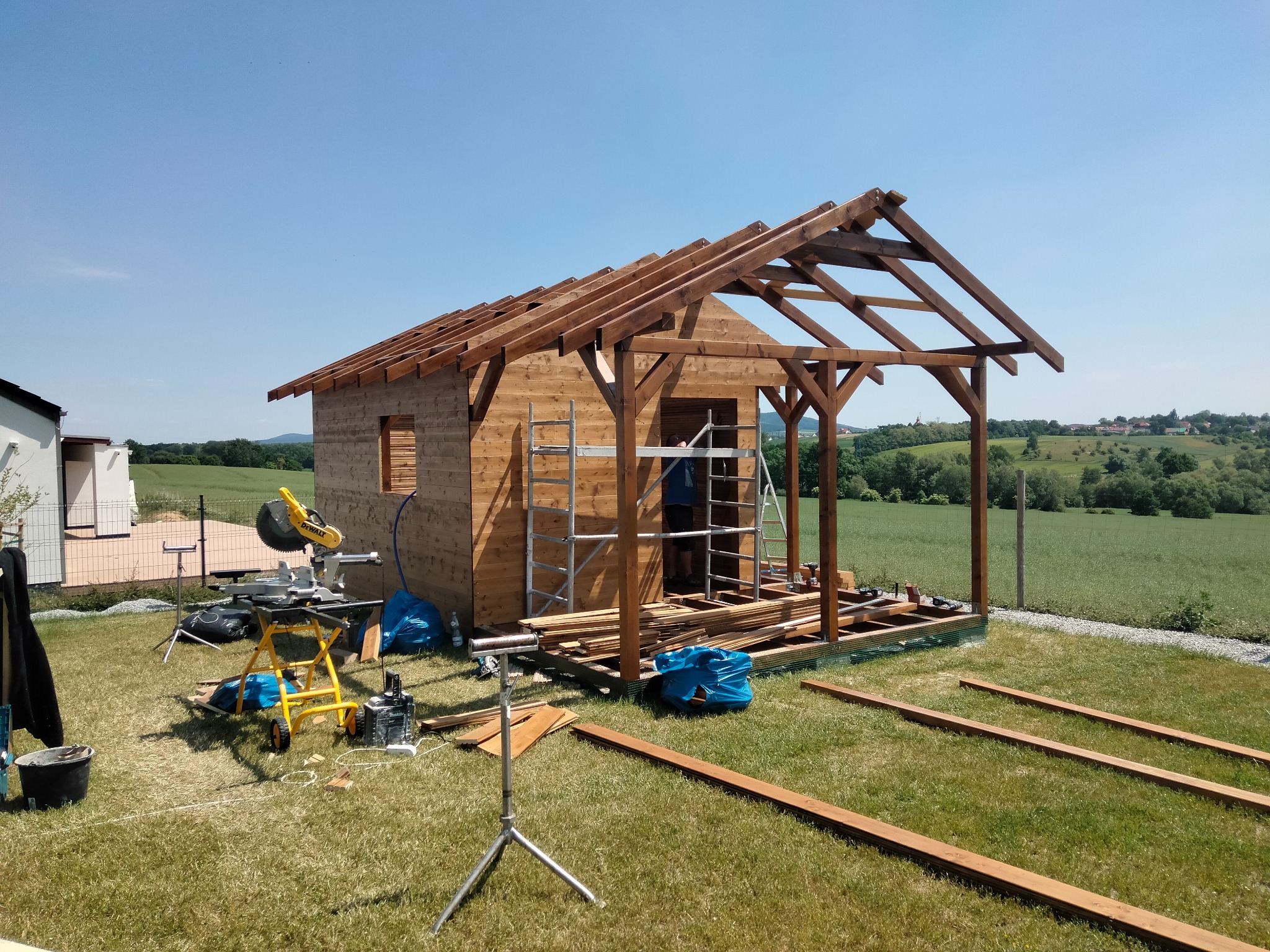 Drewniany domek letniskowy w trakcie budowy, widoczne narzędzia i elementy konstrukcyjne na tle zielonego krajobrazu. Uwagę przykuwa precyzja wykonania szkieletu.