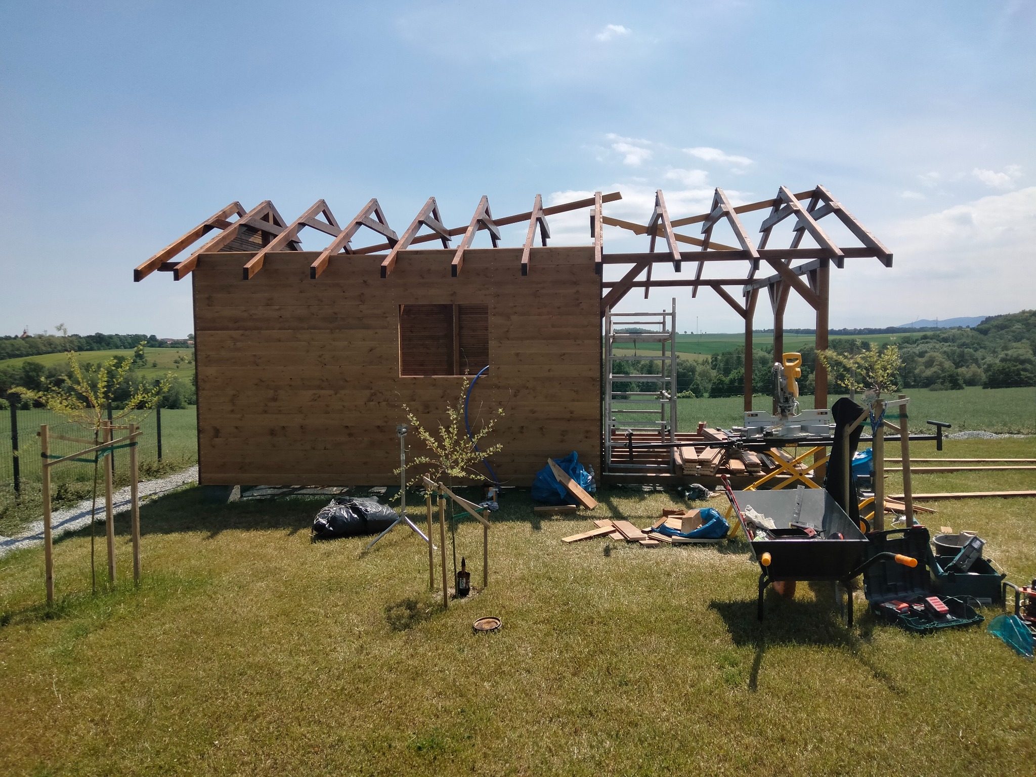 Budowa drewnianej konstrukcji z widocznym szkieletem dachu na tle zielonego krajobrazu. Wokół narzędzia i materiały budowlane.