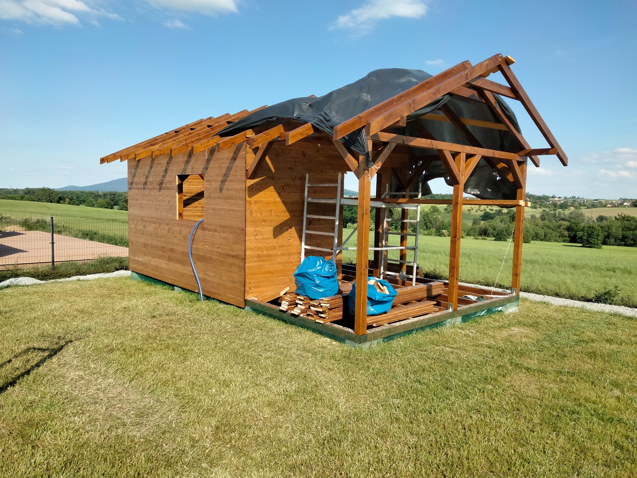 Drewniana konstrukcja domku letniskowego w trakcie budowy, widoczne elementy szkieletu i częściowo obłożone ściany, na tle zielonego krajobrazu i błękitnego nieba.