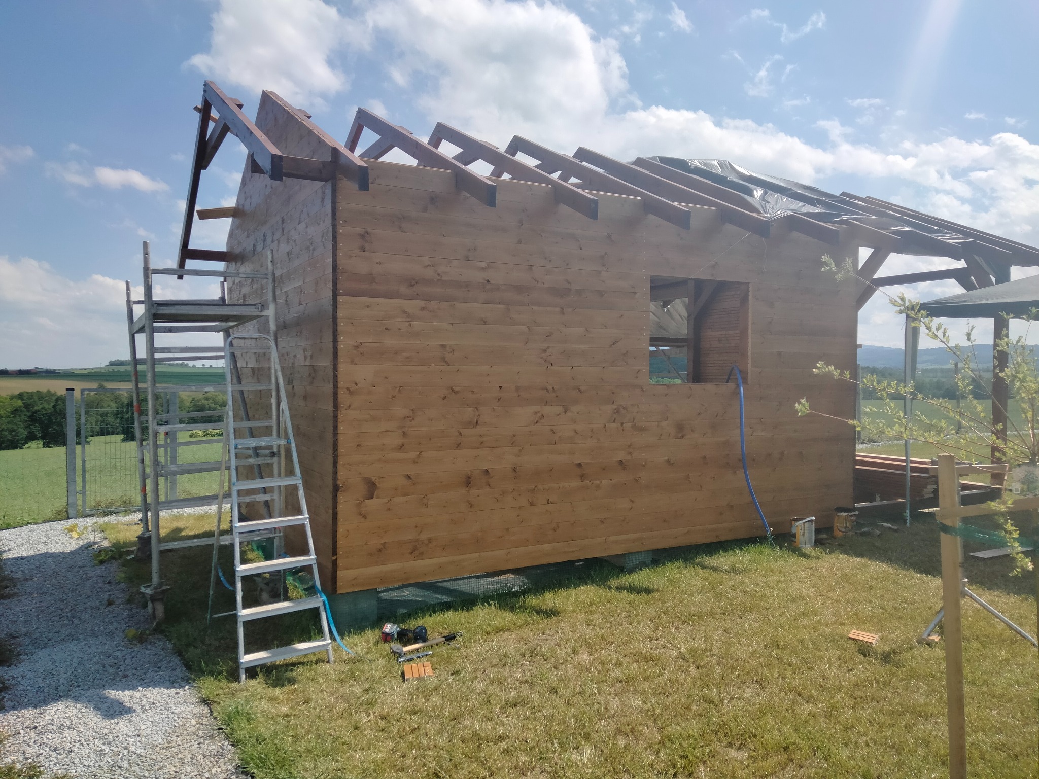 Surowy dom szkieletowy z drewnianą elewacją w trakcie budowy, z widocznym rusztowaniem i narzędziami na trawie, na tle zielonych pól i błękitnego nieba.
