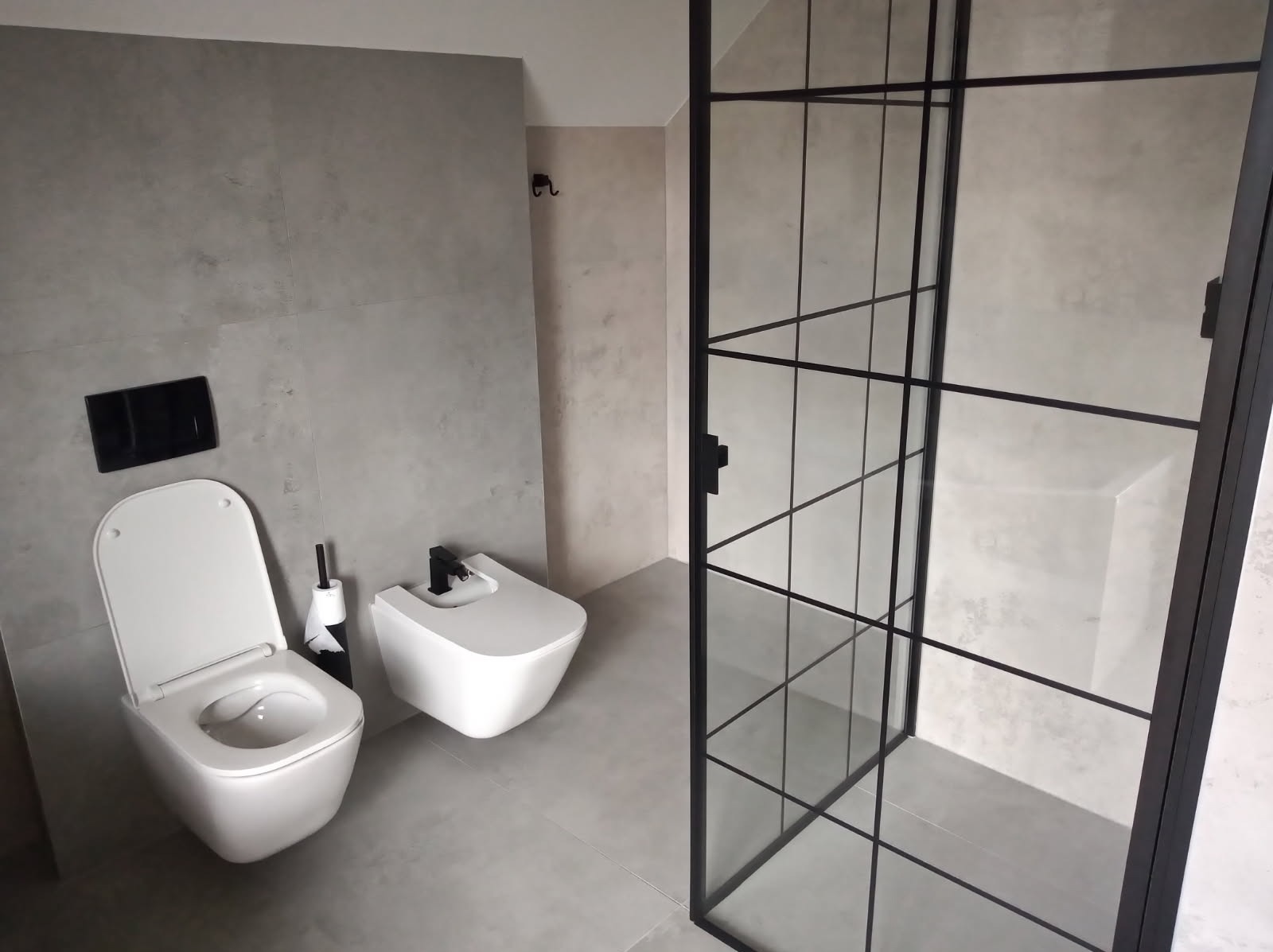 Nowoczesna łazienka w szarościach z kabiną prysznicową typu walk-in, wiszącą toaletą i bidetem. Minimalistyczny design z czarnymi akcentami.