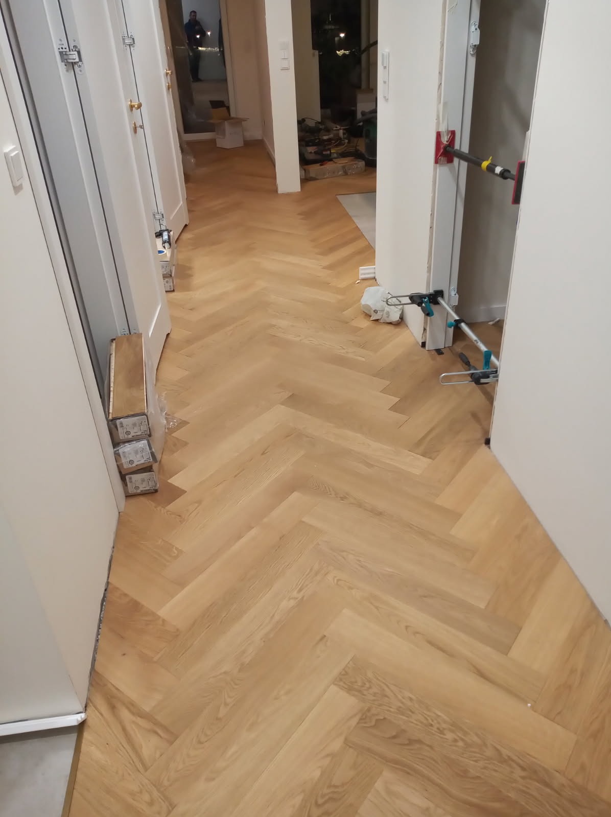 Ułożony parkiet w jodełkę w korytarzu, widoczne narzędzia do montażu drzwi, paczki paneli przy ścianie. Jasne wnętrze, neutralne kolory.