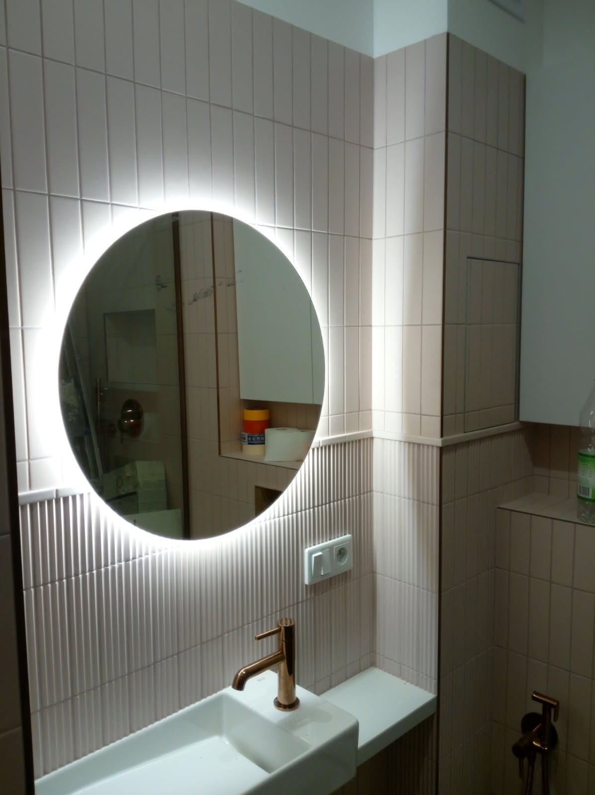 Nowoczesna łazienka z okrągłym lustrem LED, beżowe płytki z delikatnym wzorem, biała umywalka z miedzianą baterią. Widoczny fragment instalacji prysznicowej.