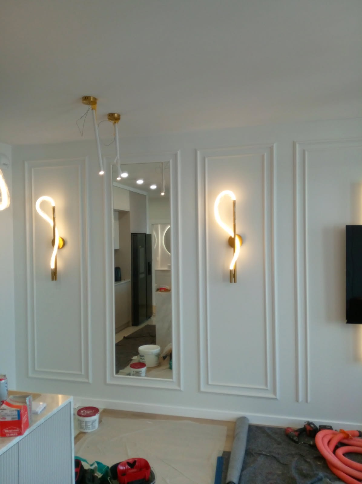 Białe ściany z dekoracyjnymi listwami i oświetleniem LED. Widoczne narzędzia malarskie i wiadro z farbą. Lustro w ozdobnej ramie odbija wnętrze pomieszczenia.