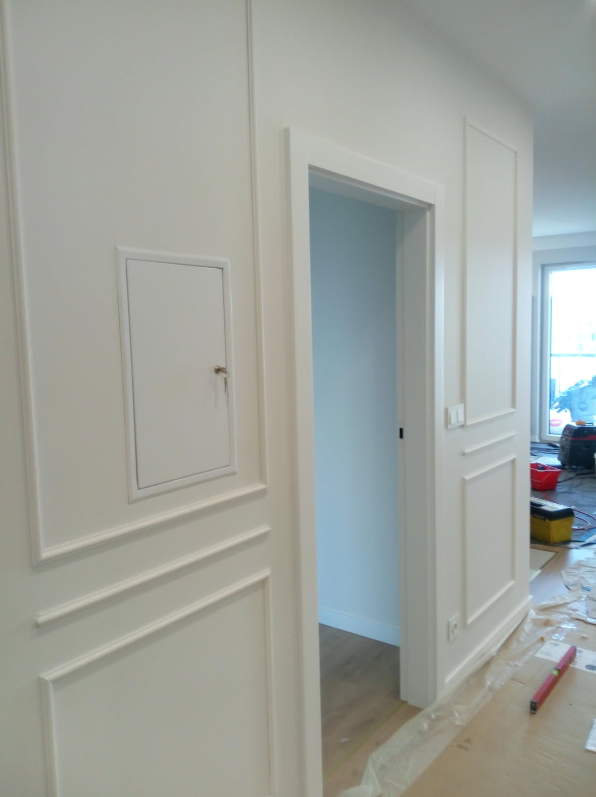 Białe, klasyczne wykończenie ściany z dekoracyjnymi listwami i ukrytymi drzwiczkami rewizyjnymi, widok na otwarte drzwi i fragment pomieszczenia w trakcie prac wykończeniowych.