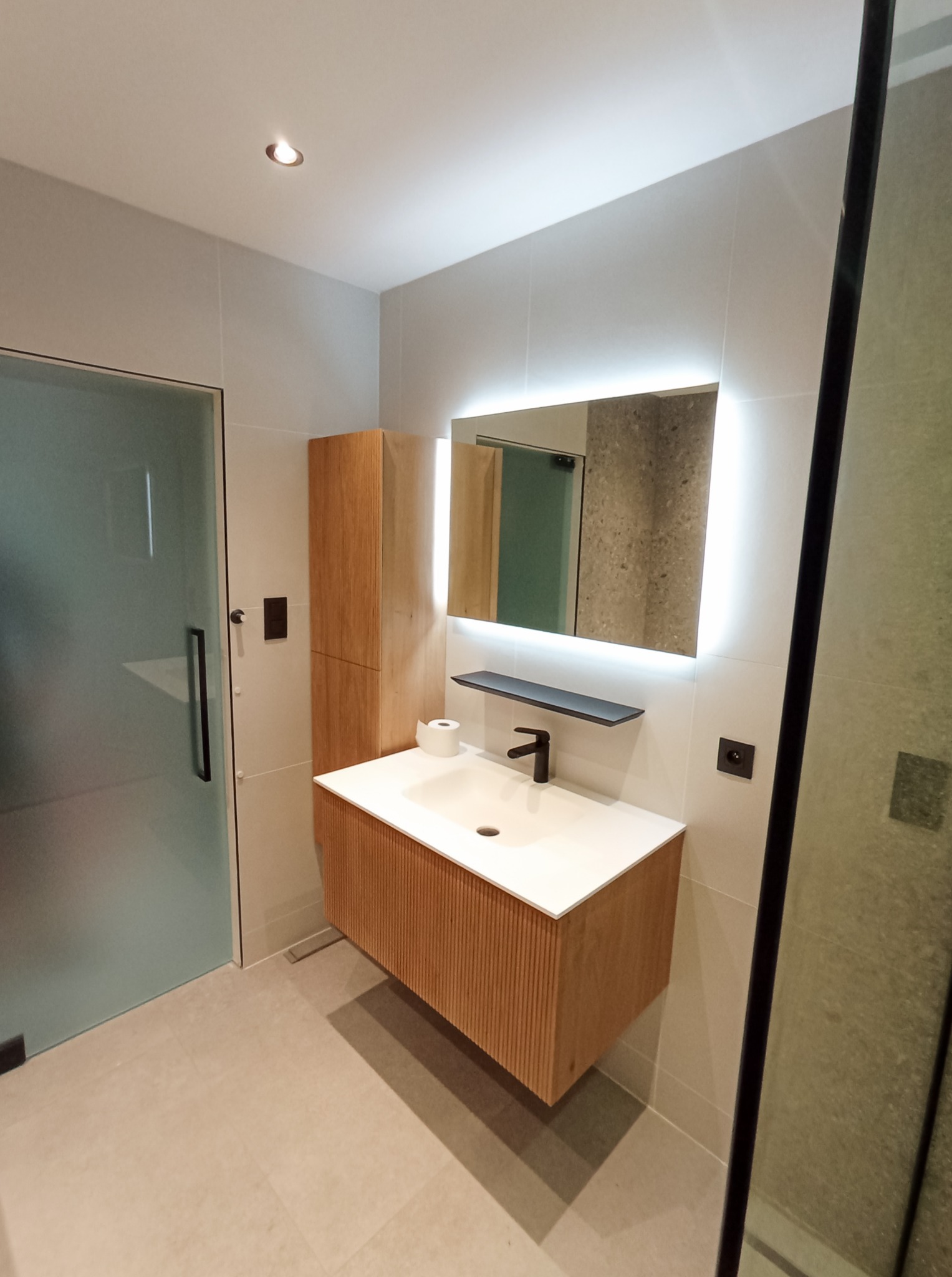 Nowoczesna łazienka z drewnianą szafką pod umywalką, podświetlanym lustrem i kabiną prysznicową z czarnymi ramami. Minimalistyczny design i jasne płytki.