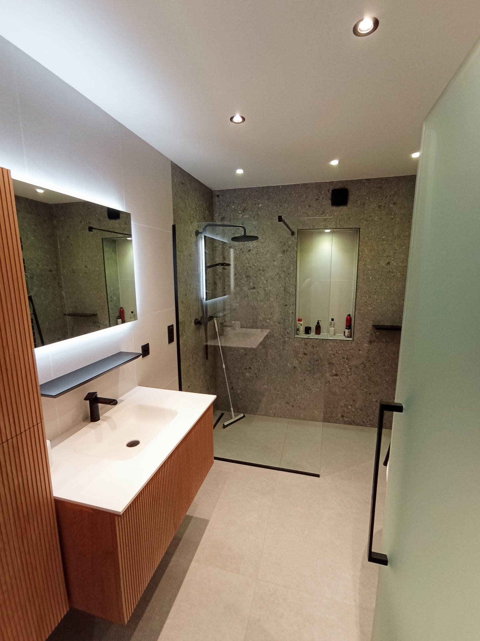 Nowoczesna łazienka z kabiną prysznicową typu walk-in, umywalką z szafką z lamelami i podświetlanym lustrem. Szare płytki na ścianach i beżowa podłoga.