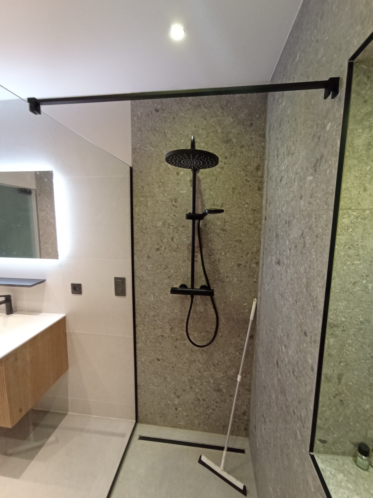 Nowoczesna kabina prysznicowa walk-in z czarną armaturą i szklaną ścianką. Minimalistyczny design, szare płytki imitujące kamień, oświetlenie LED lustra.