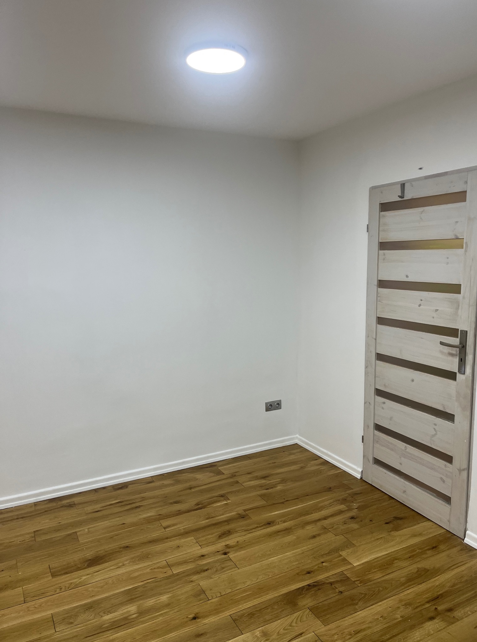 Jasne panele podłogowe w nowym pokoju z białą ścianą i drzwiami z przeszkleniami. Widoczne gniazdko elektryczne i okrągła lampa sufitowa.
