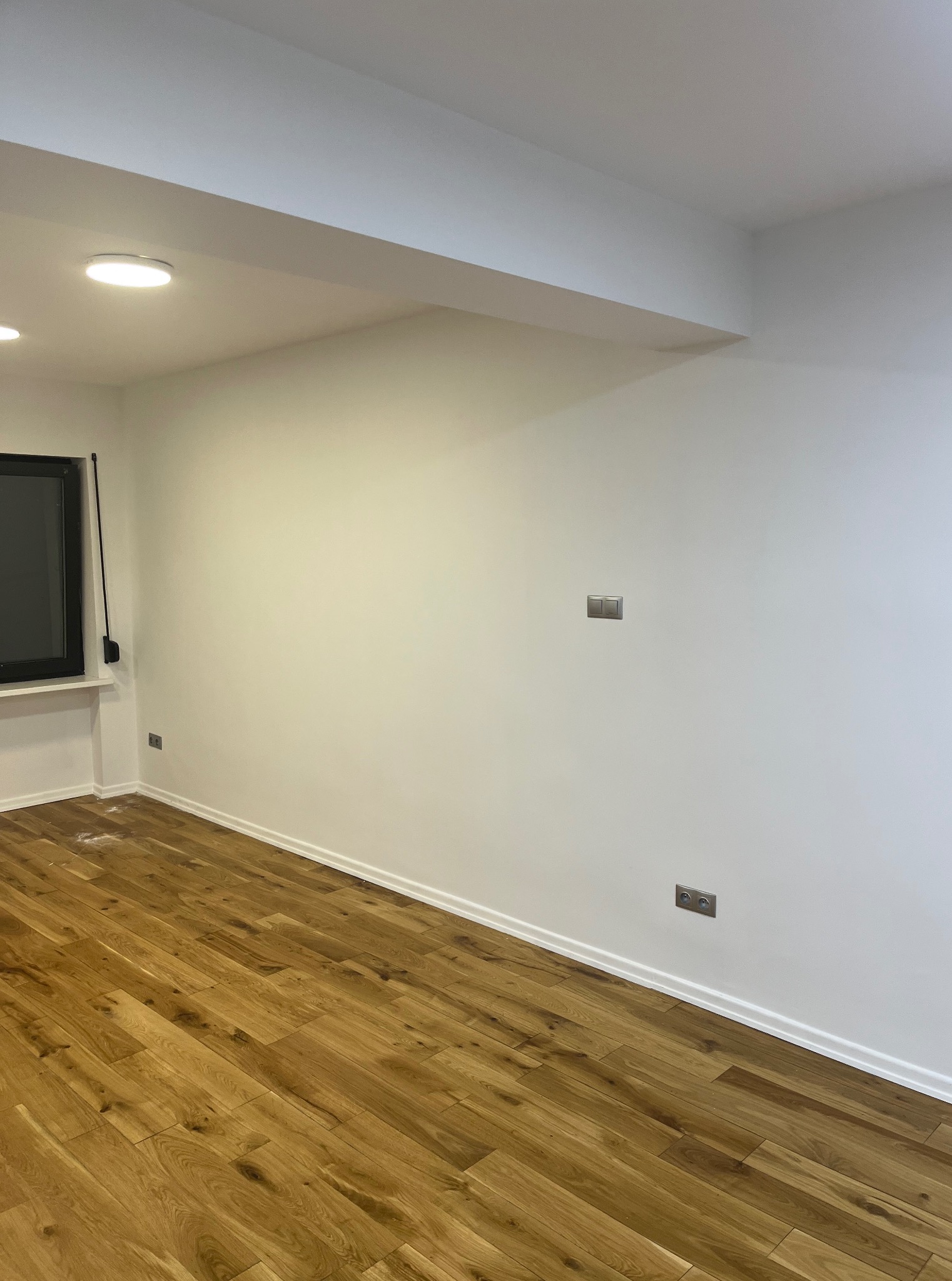 Jasne panele podłogowe ułożone w pokoju z białymi ścianami i listwami przypodłogowymi. Widoczne gniazdka elektryczne i włącznik światła.