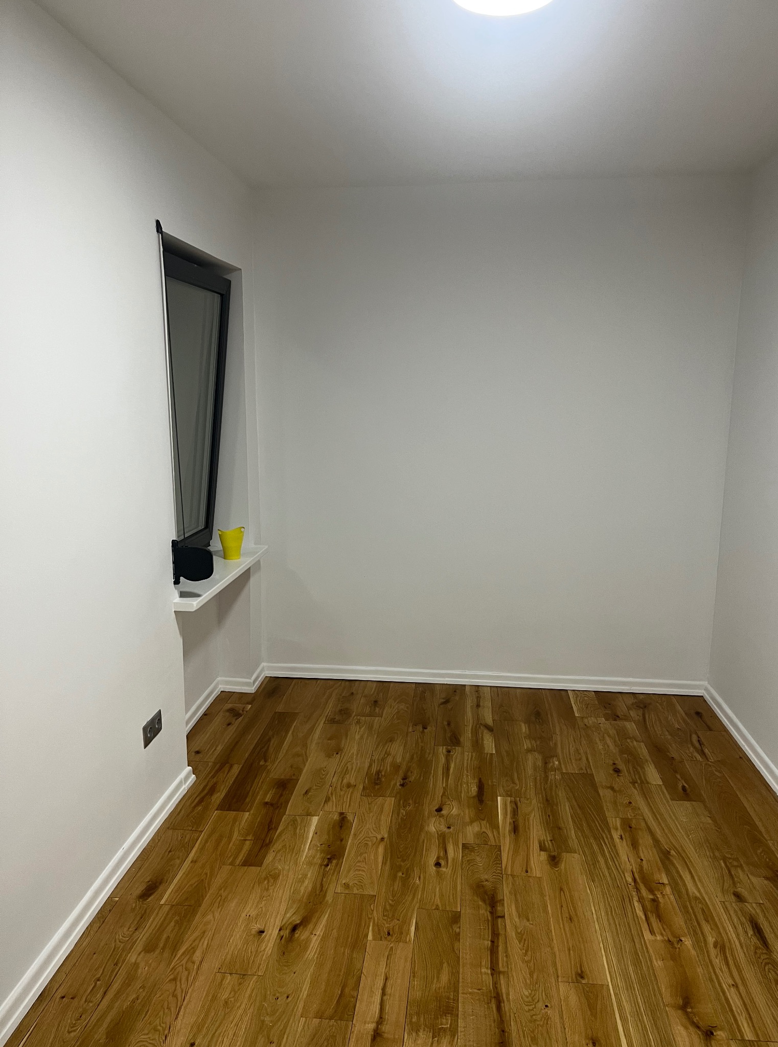 Jasne, puste pomieszczenie po remoncie z drewnianą podłogą, białymi ścianami i oknem z czarną ramą. Na parapecie stoi żółty kubek. Widoczne listwy przypodłogowe.