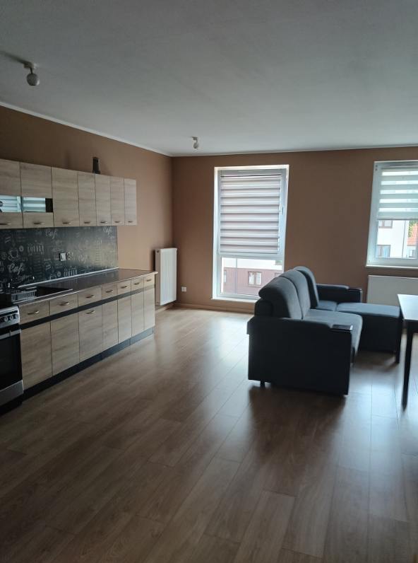 Jasne panele podłogowe w salonie z aneksem kuchennym, brązowe ściany, szara sofa i okna z roletami. Widok na wykończone wnętrze mieszkania.