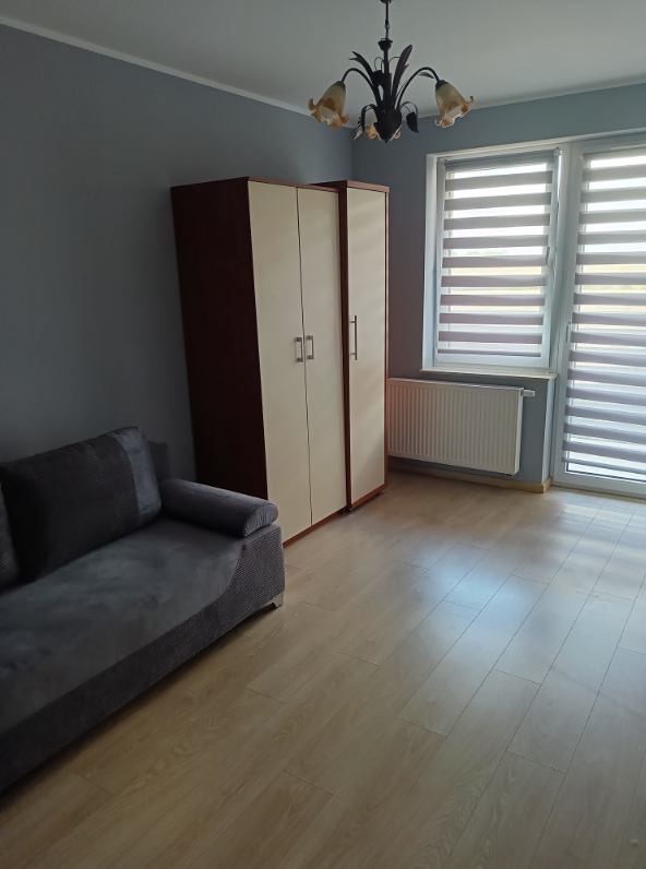 Pokój po odświeżeniu: szara ściana, drewniana podłoga, sofa i szafa w odcieniach brązu i beżu. Okno z żaluzjami i dekoracyjny żyrandol.