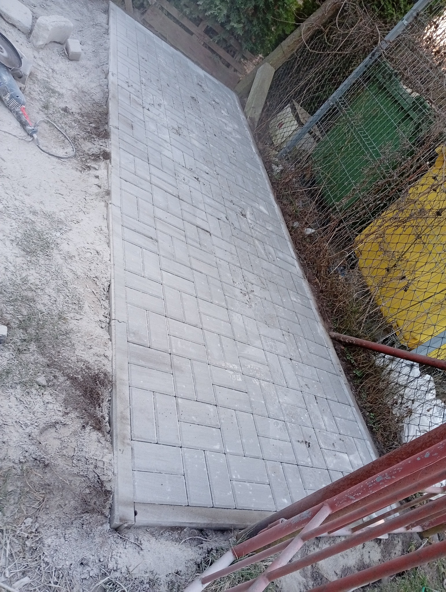 Ułożona szara kostka brukowa na podwórku, widoczna z góry pod kątem. Obok ziemia i trawa, w tle siatka ogrodzeniowa i zieleń. Częściowo widoczna szlifierka kątowa.
