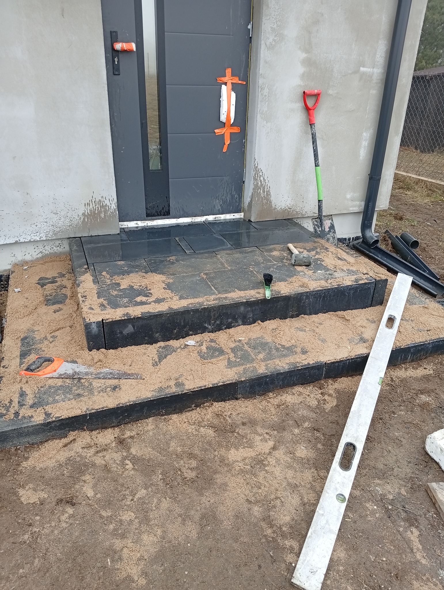 Nowe, czarne schody wejściowe z płytek, częściowo zasypane piaskiem. Widoczne narzędzia: poziomica, piła ręczna, szpachelka. Drzwi wejściowe zabezpieczone taśmą.