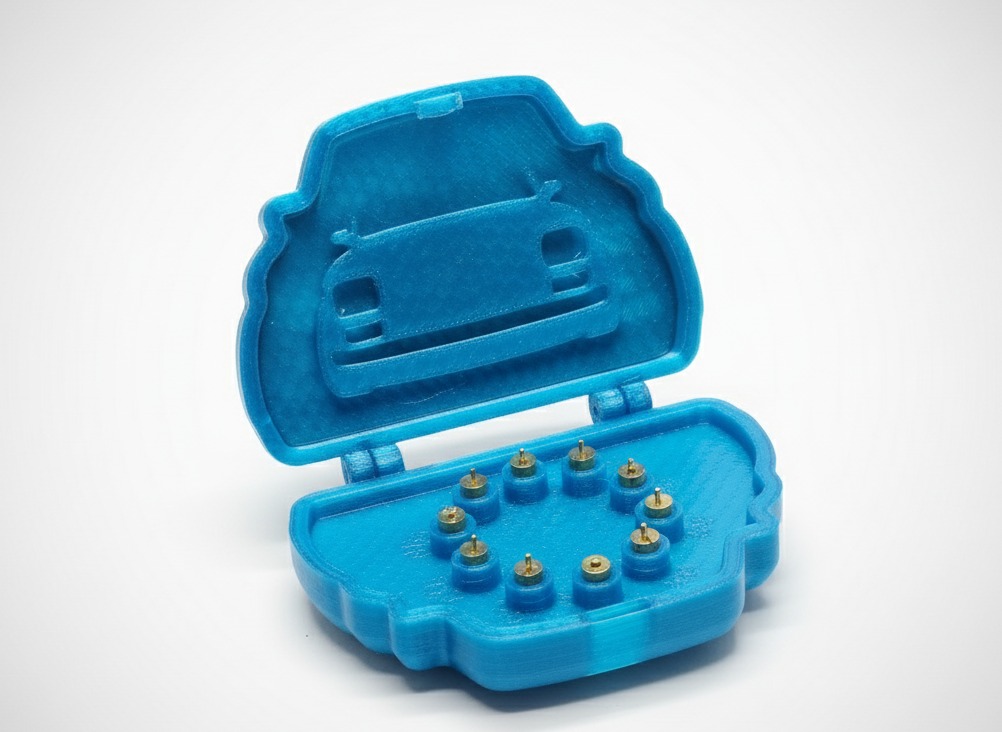 Niebieskie pudełko z tworzywa sztucznego, wydrukowane w 3D, otwarte, z wytłoczonym kształtem samochodu na pokrywie i metalowymi elementami wewnątrz. Widoczne ślady druku 3D.