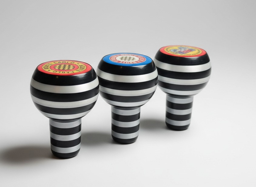 Trzy czarno-srebrne gałki zmiany biegów z różnymi kolorowymi emblematami na górze, ustawione w rzędzie na białym tle, oświetlone z góry.