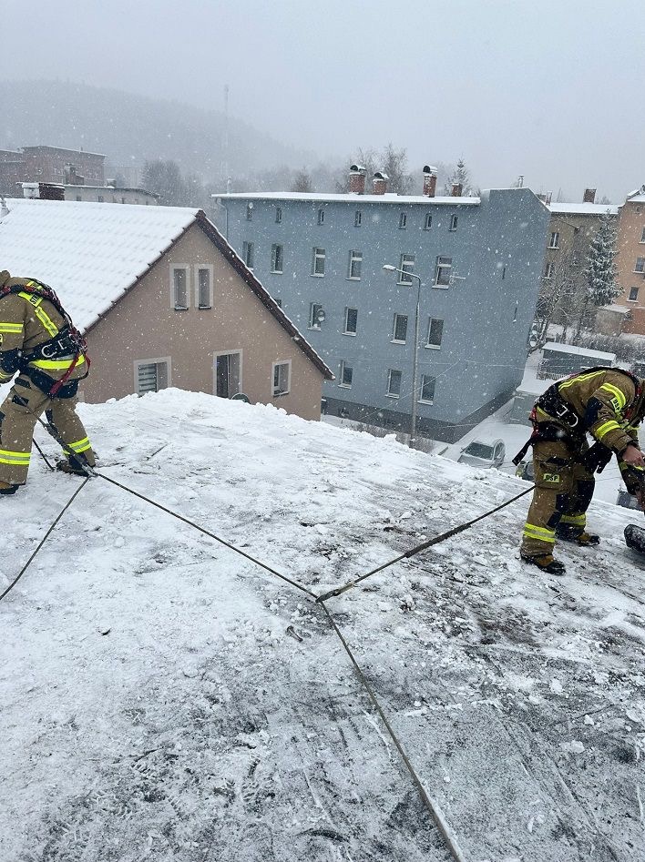 Strażacy zabezpieczeni linami pracują na ośnieżonym dachu budynku podczas opadów śniegu. W tle zabudowa miejska i wzgórze.