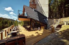 Nowoczesny budynek z ciemnoszarym dachem dwuspadowym, drewnianym tarasem i szklanymi ścianami, otoczony lasem iglastym w słoneczny dzień. Nietypowa architektura.