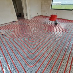 C.O InstalGreen Adam Ptak - Czerwone rury ogrzewania podłogowego ułożone na srebrnej folii z czarną siatką w surowym wnętrzu z oknem i rolką rur na wózku. Widok z góry.