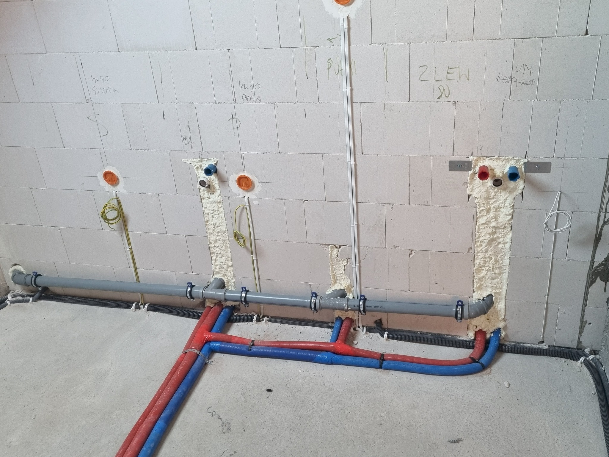 Instalacja wodno-kanalizacyjna w surowym stanie: rury PEX (czerwone i niebieskie) prowadzone wzdłuż ściany z betonu komórkowego, widoczne punkty przyłączeniowe.