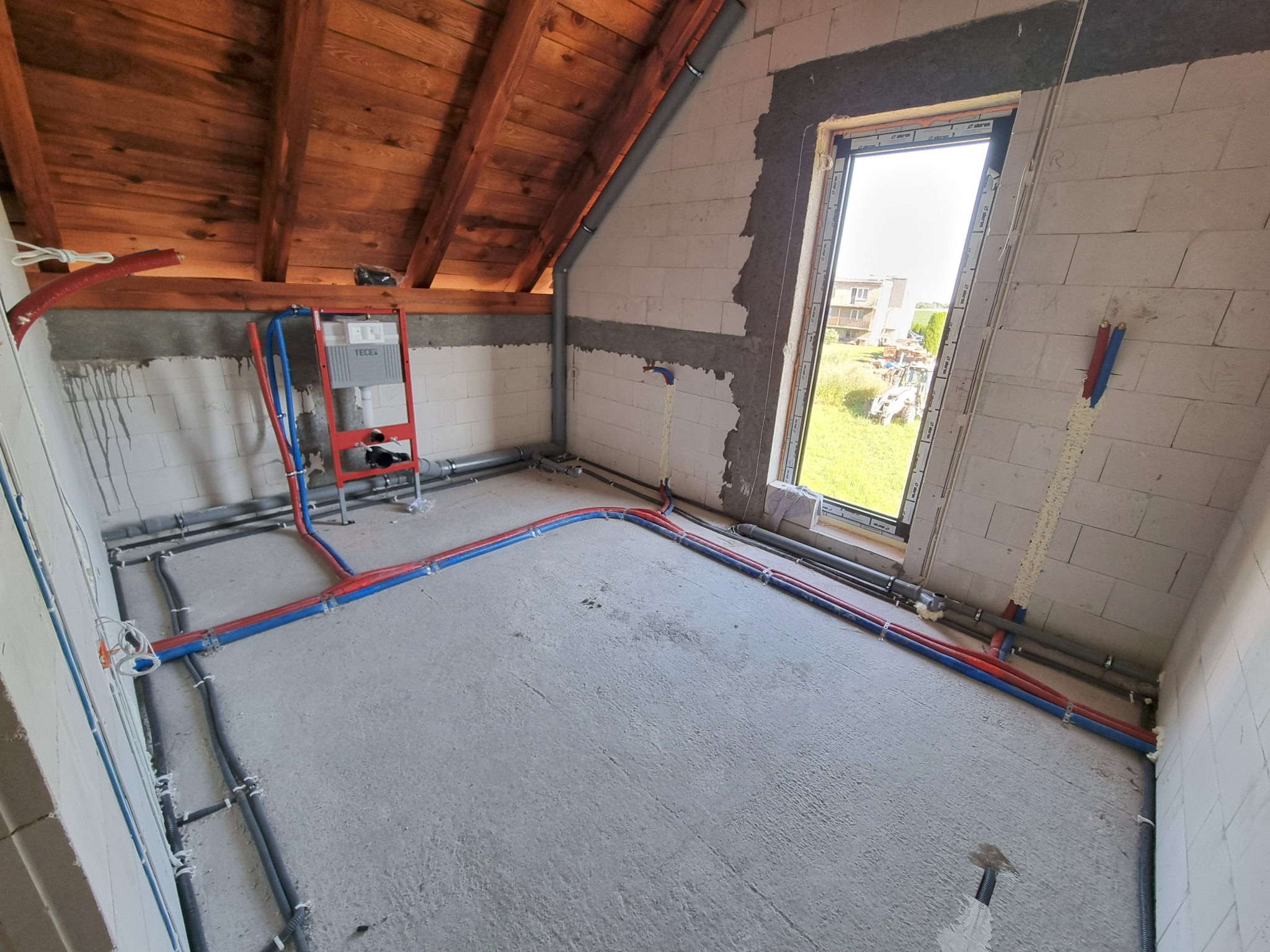 Instalacja wodna i kanalizacyjna w surowym wnętrzu poddasza z oknem. Rury w kolorze niebieskim i czerwonym prowadzone po podłodze i ścianach. Widoczny stelaż podtynkowy WC.