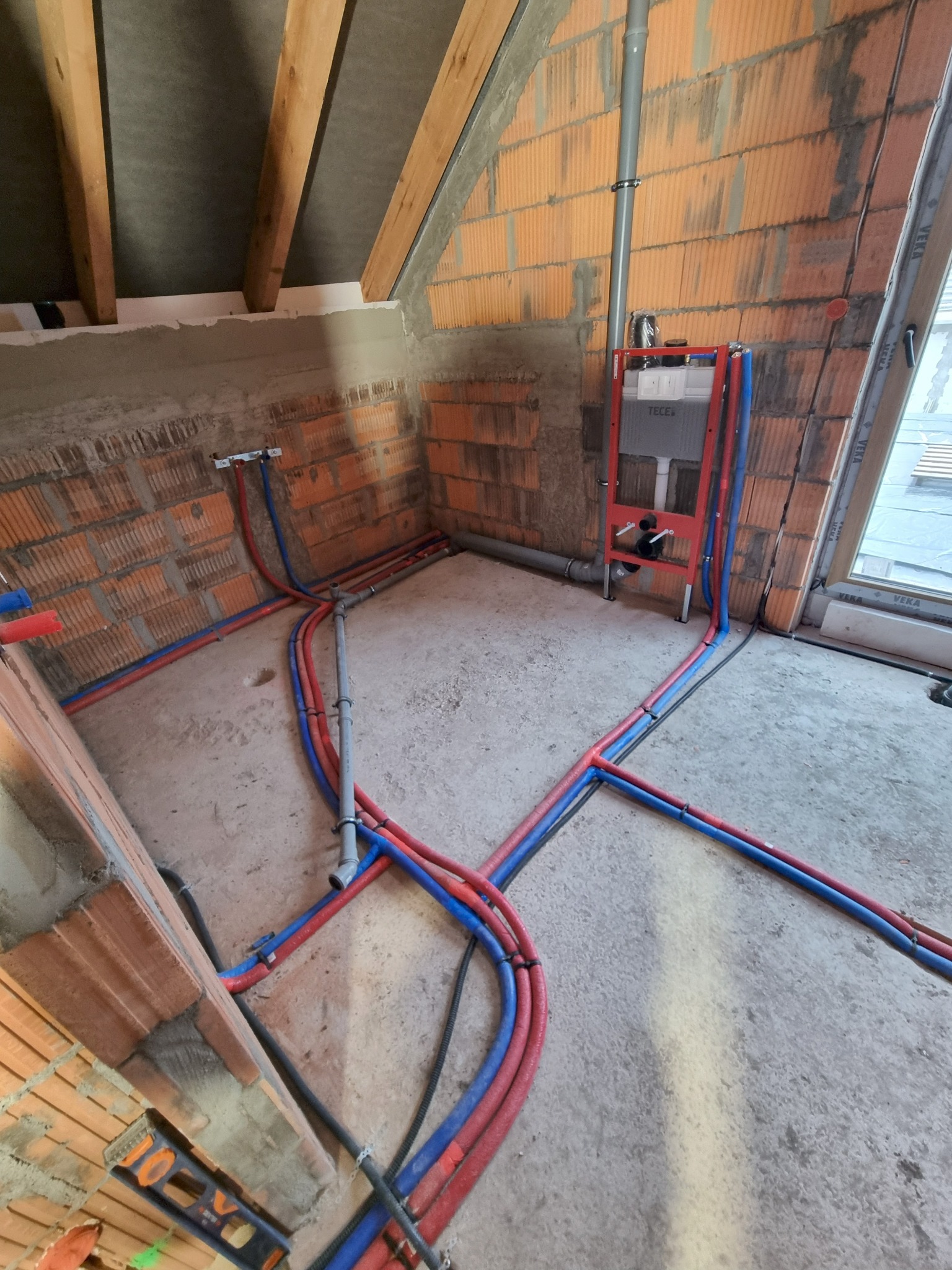 Instalacja wodna i kanalizacyjna w surowym wnętrzu poddasza. Czerwone i niebieskie rury prowadzone po podłodze, widoczny stelaż podtynkowy WC i fragment okna.
